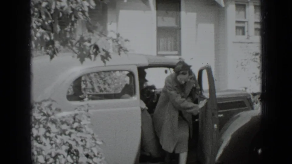 In 1939 New York home-movie footage, a woman in a long coat steps from a parked vintage car near a house.