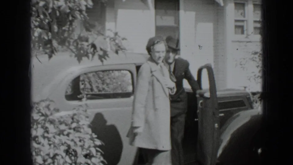A woman in a coat and a suited man stand near an open car door outside a New York house in 1939.