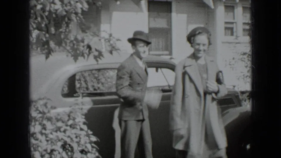 The couple smiles beside the parked car, with the house and leafy shrubs visible behind them.