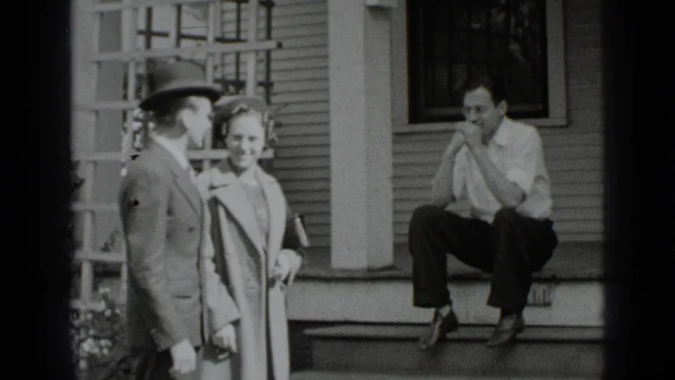 The couple approaches the house steps while another man sits nearby, watching from the porch area.