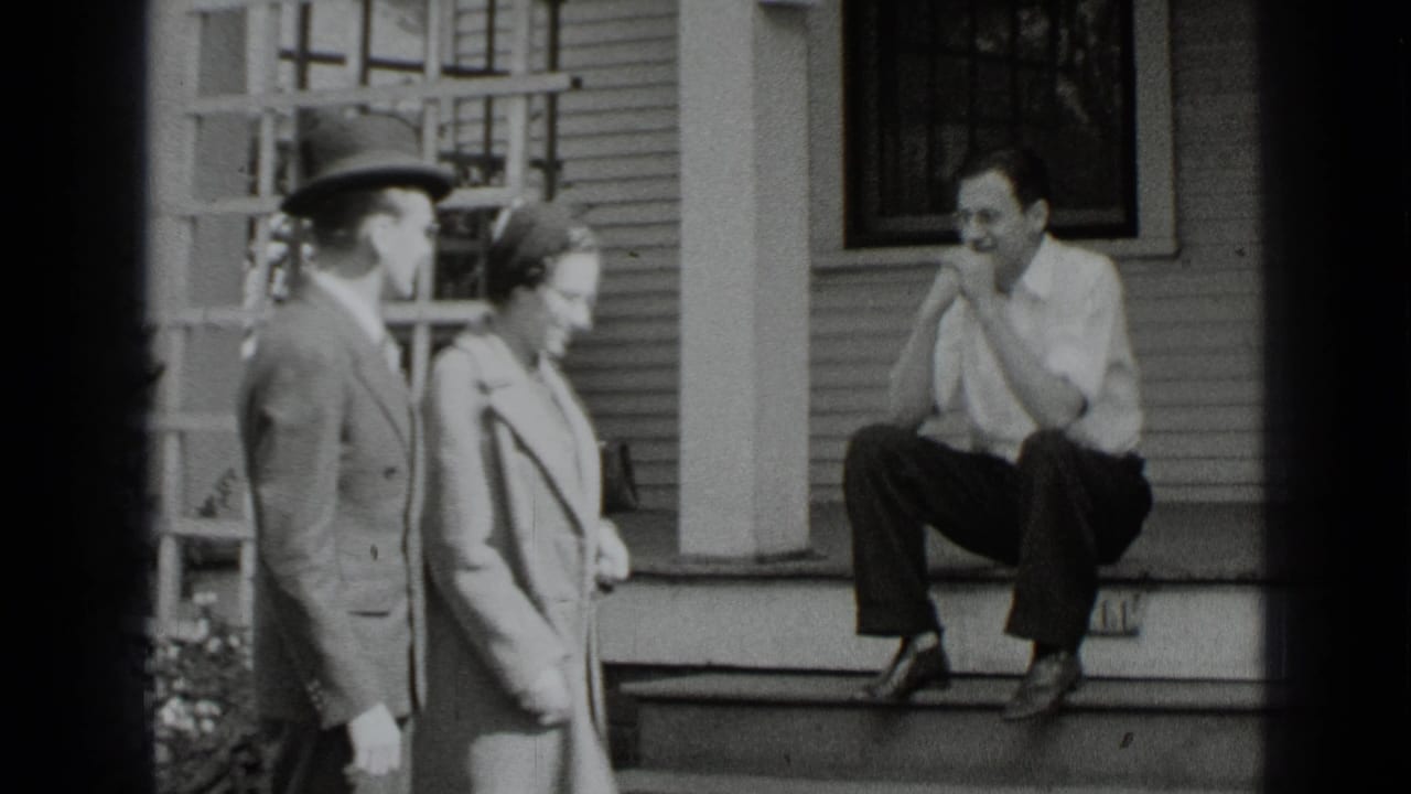 In 1939 New York home-movie footage, a suited man and a woman in a coat stand near porch steps as a man sits outside the house.