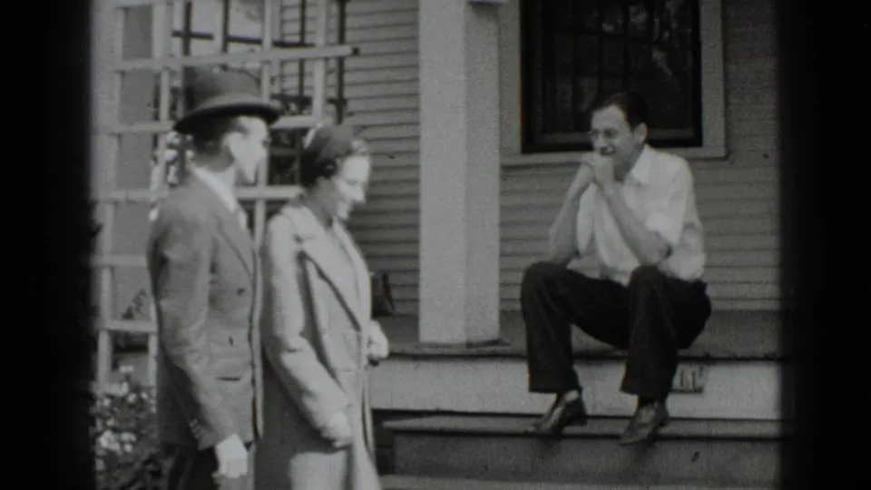 In 1939 New York home-movie footage, a suited man and a woman in a coat stand near porch steps as a man sits outside the house.