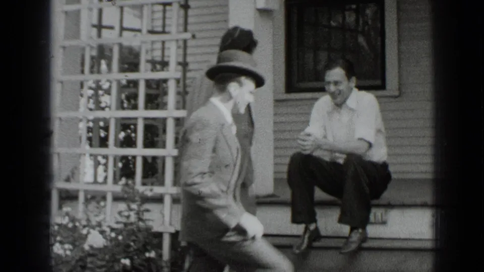 A man in a suit and hat moves across the frame while a seated man smiles on the porch of a New York house.