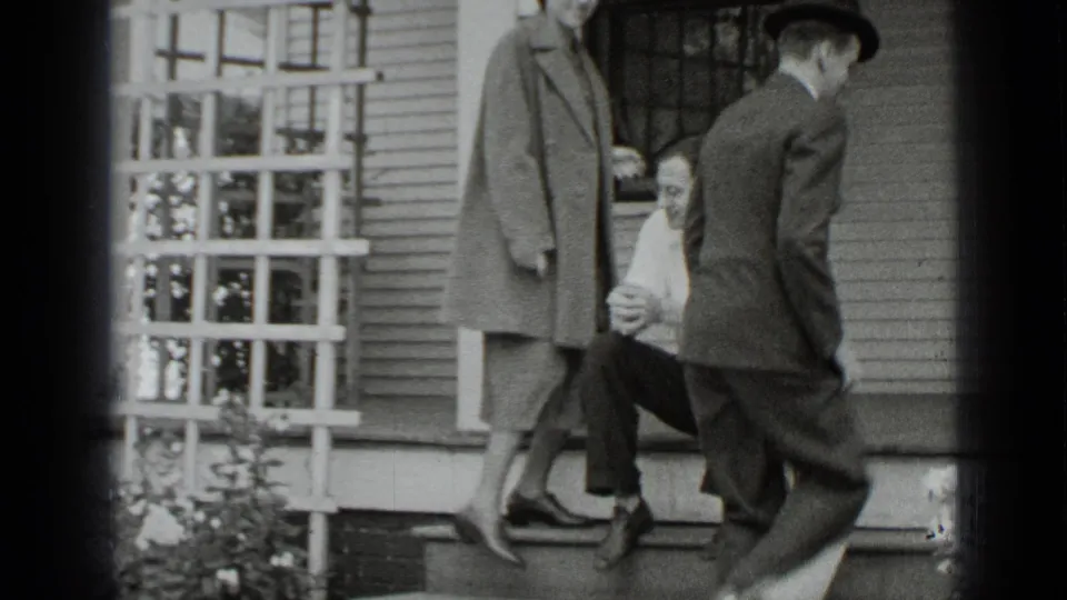 The woman in a coat and the suited man stand near the steps, facing a seated man outside a New York house.