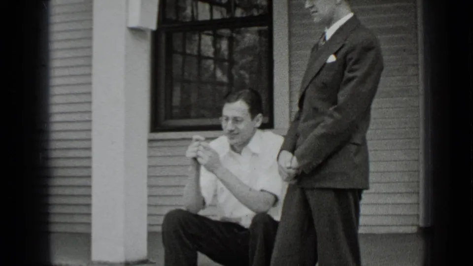 A seated man in a light shirt smiles and gestures while a suited man stands nearby on the porch of a New York house.