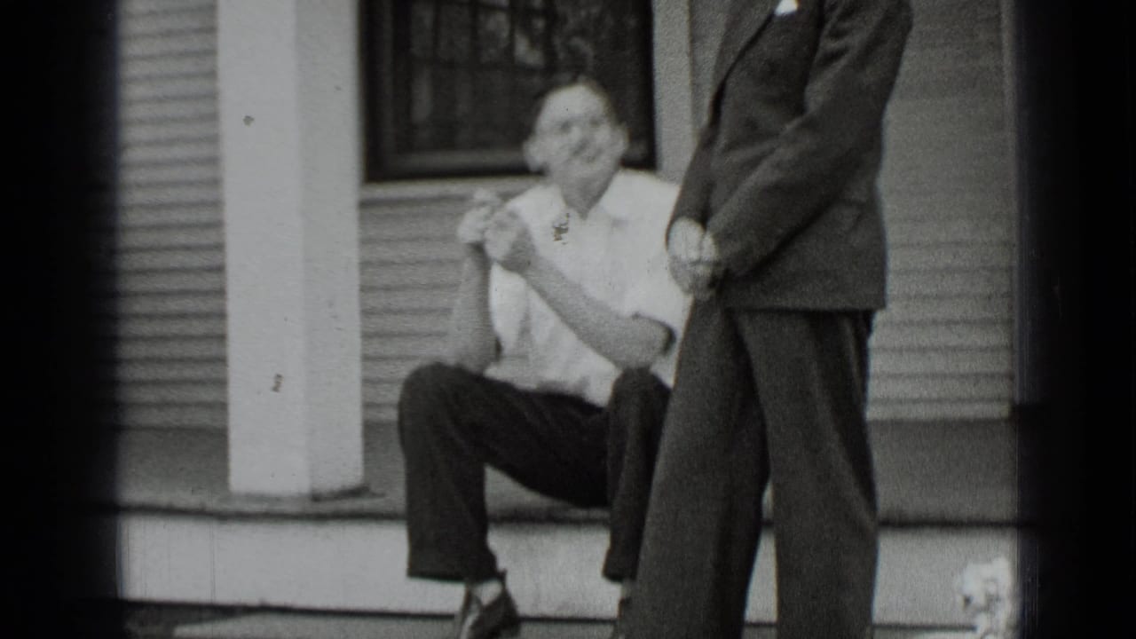 Man seated on porch steps while another suited man stands nearby.