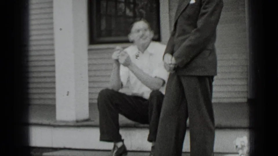 1939 New York home-movie frame showing a man seated on porch steps beside a standing man in a suit outside a clapboard house.