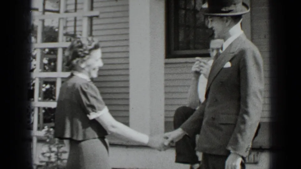The woman and suited man complete a handshake near the porch, with house siding and a window behind them in this 1939 New York home-movie frame.
