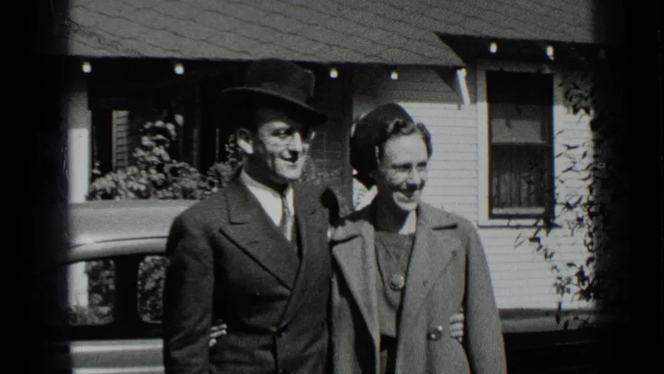 A smiling couple stands arm-in-arm outside a New York house, with a parked car and porch visible in the 1939 home-movie frame.