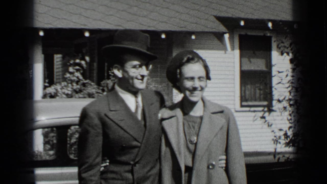 In 1939 New York home-movie footage, an adult couple in formal coats and hats smiles while posing outside a residential house.