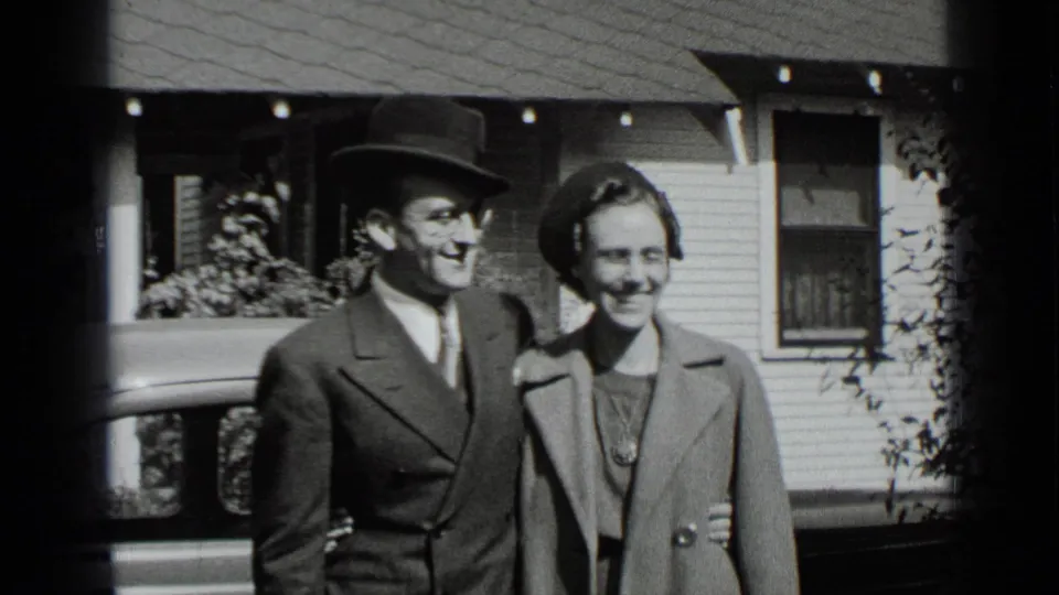 In 1939 New York home-movie footage, an adult couple in formal coats and hats smiles while posing outside a residential house.