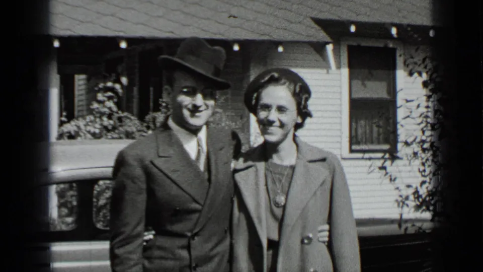 The couple stands arm in arm outside a New York residence, with 1930s clothing and eyeglasses visible in the black-and-white frame.