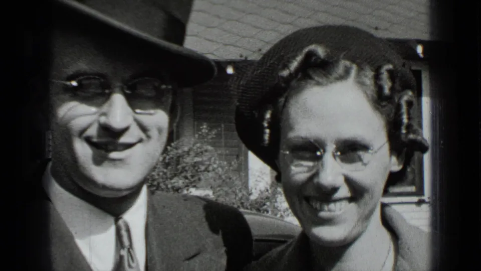 A close 1939 home-movie frame shows the man in a fedora and the woman in round glasses smiling near a residential exterior.