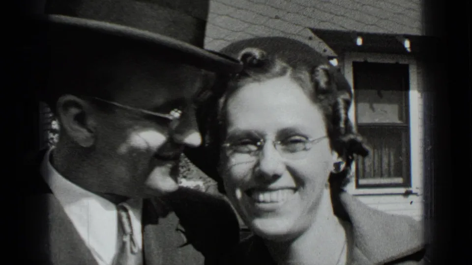 The man leans affectionately toward the smiling woman in a close New York home-movie shot, with a house window visible behind them.