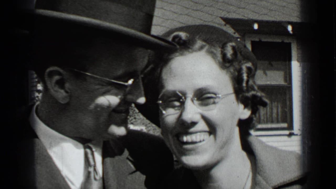 Smiling man in a fedora and woman with glasses standing close to the camera outside.