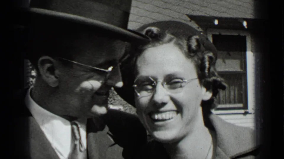 In a 1939 New York home-movie frame, a smiling couple stands close together outdoors, with the man in a fedora and the woman wearing glasses.