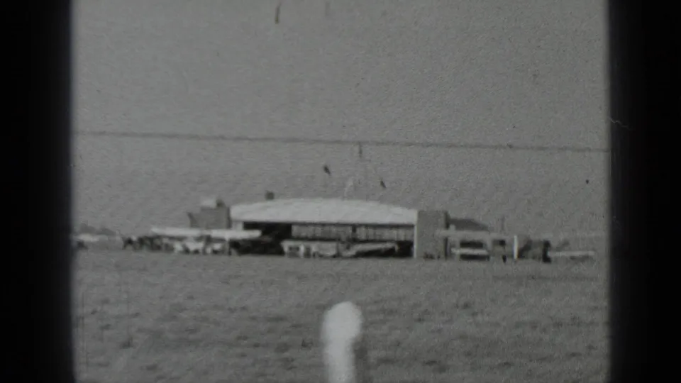 A distant airfield view in New York shows a low hangar-like building, small aircraft, and open ground, with a finger briefly visible near the lens.