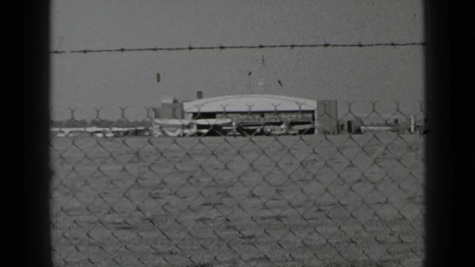 The airfield appears through a chain-link fence, with a low hangar-like structure and small aircraft visible beyond the fenced foreground.