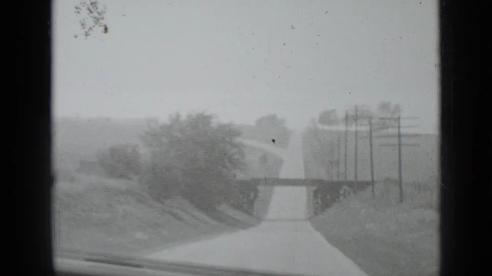 A moving-vehicle view shows a road, utility poles, roadside fencing, and soft landscape detail, likely near the airfield sequence described in the metadata.
