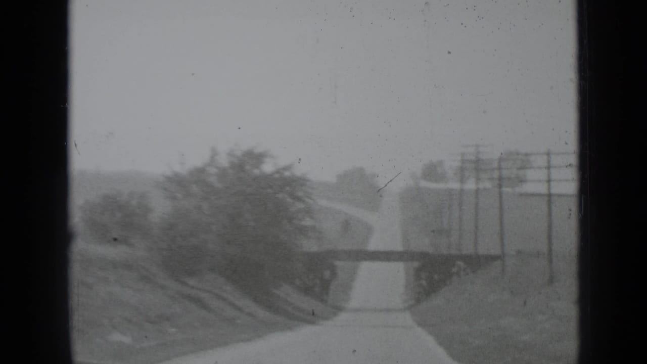 Narrow rural road leading toward a low bridge with trees and utility poles nearby.