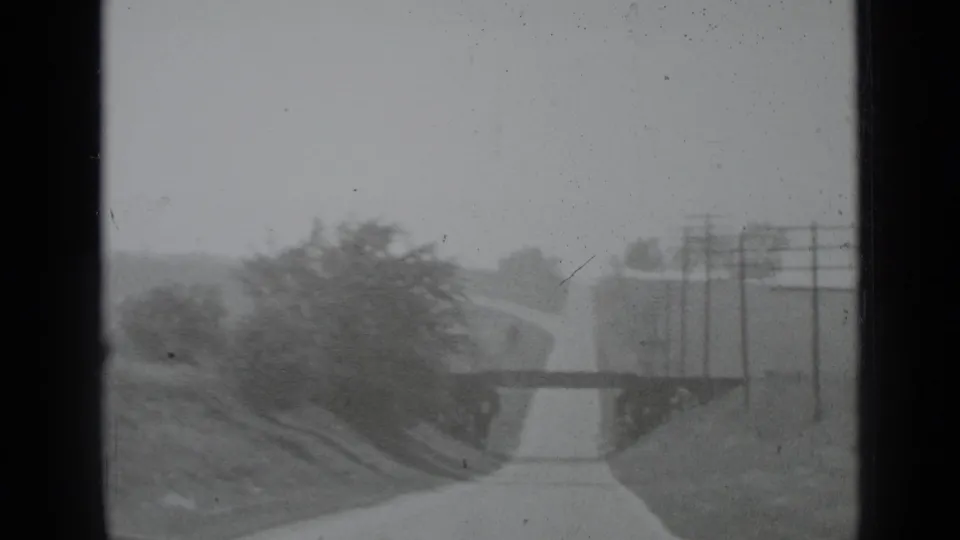 1939 New York home-movie frame showing a narrow road approaching a small bridge, with trees and utility poles along the roadside.
