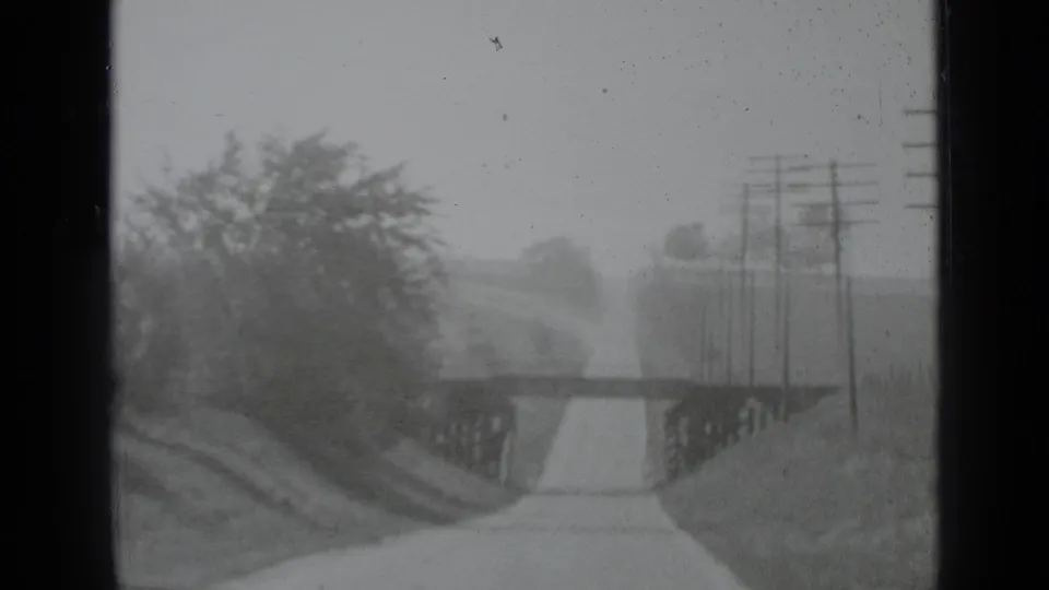 A soft black-and-white 8mm view of a rural New York road nearing a low bridge, with utility poles standing beside the route.