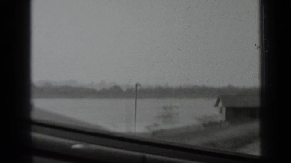 A soft black-and-white view looks past dark interior framing toward open water, a shoreline, and a small structure at the right.