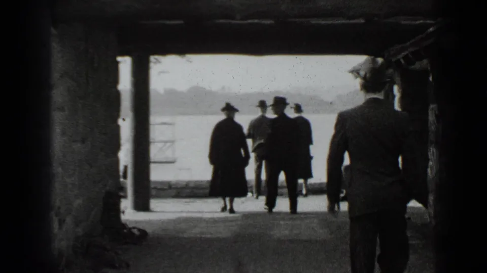 In 1939 New York home-movie footage, several people in coats and hats walk through a covered waterfront passage toward bright open water.