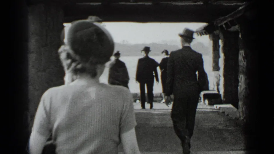 A woman in a rounded hat is seen from behind while other suited pedestrians continue toward the waterfront in black-and-white 8mm footage.