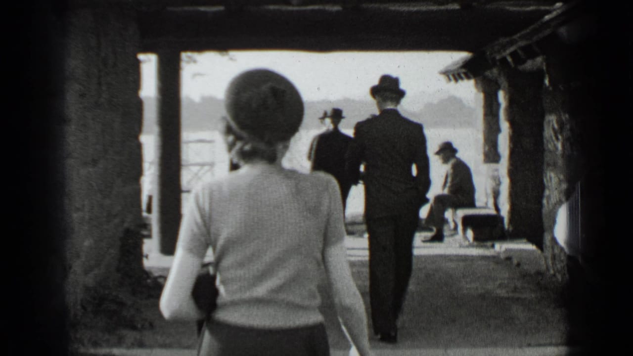 Woman in hat walks through a covered lakeside structure toward people near the water.