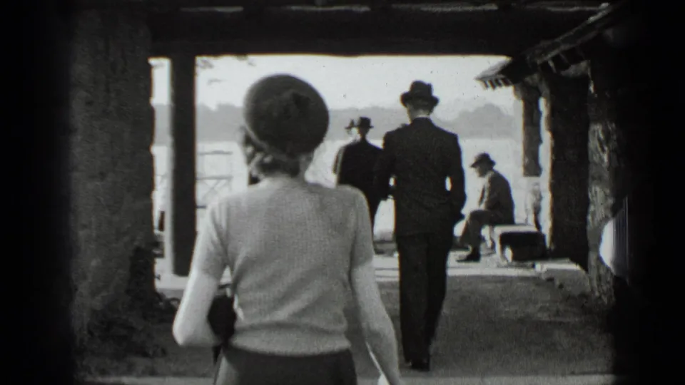 1939 New York home-movie frame showing a woman in period dress walking beneath a shaded lakeside shelter toward suited figures by the water.