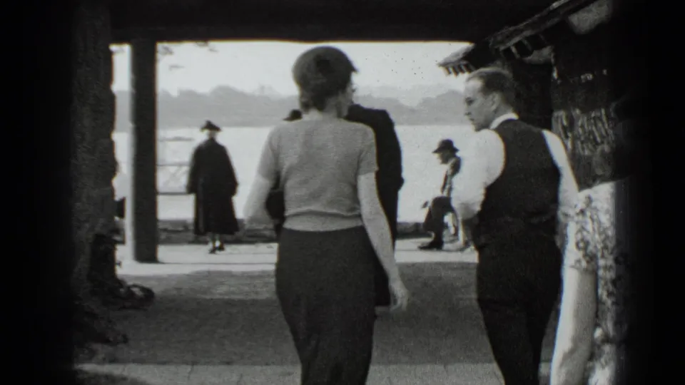 1939 New York lakeside scene with a woman and a man in a vest walking under a shelter while other visitors gather near the water.
