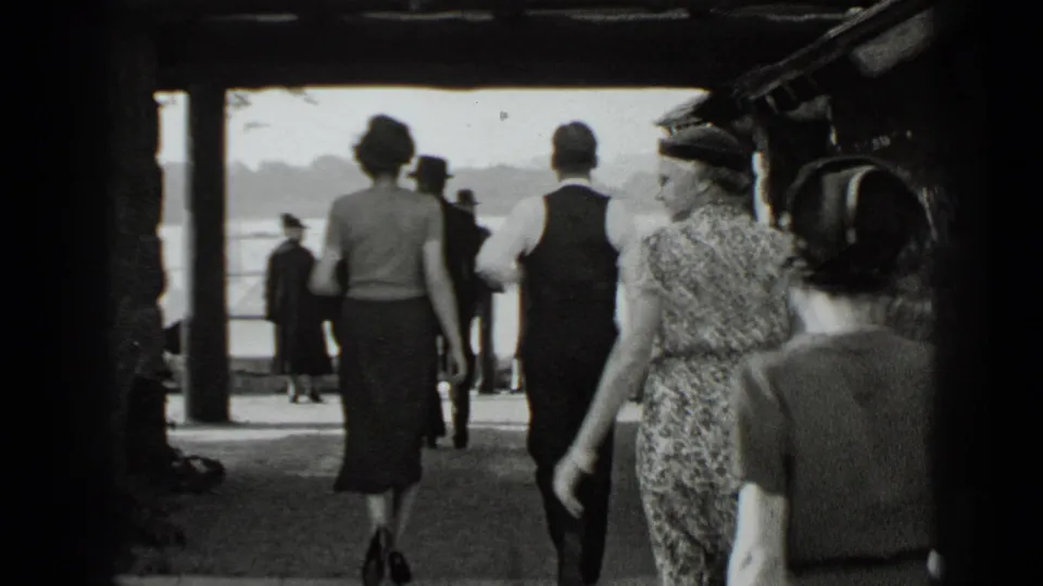 1939 New York waterfront frame showing several adults in dresses, hats, and formal clothing walking toward the bright lakeside opening.