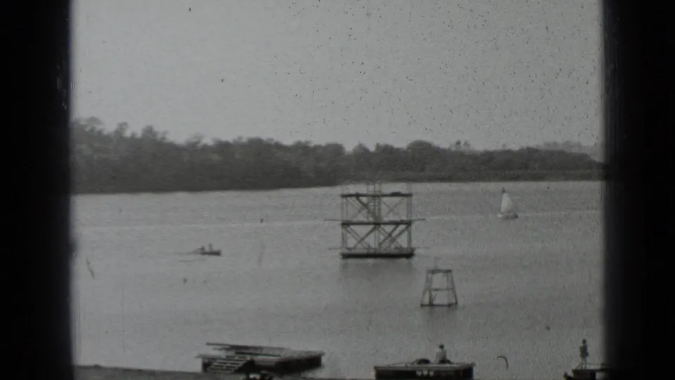 1939 New York home-movie view across a lake with small boats, a sailboat, dock platforms, and a wooded distant shoreline.
