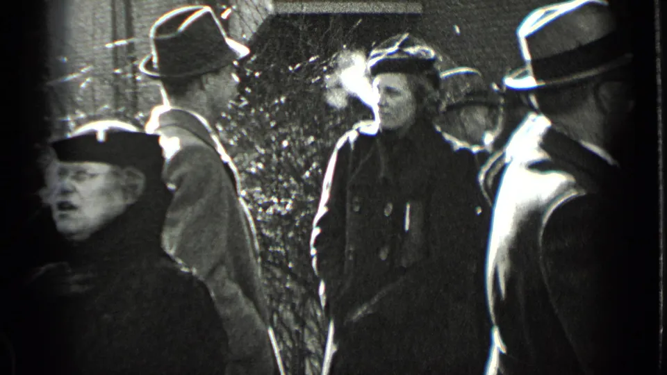 In 1939 New York, bundled adults in coats and hats socialize on a sidewalk as cold breath is visible in the winter air.