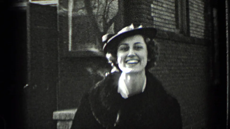 A smiling woman in a dark winter coat and hat looks toward the home-movie camera on a New York sidewalk in 1939.