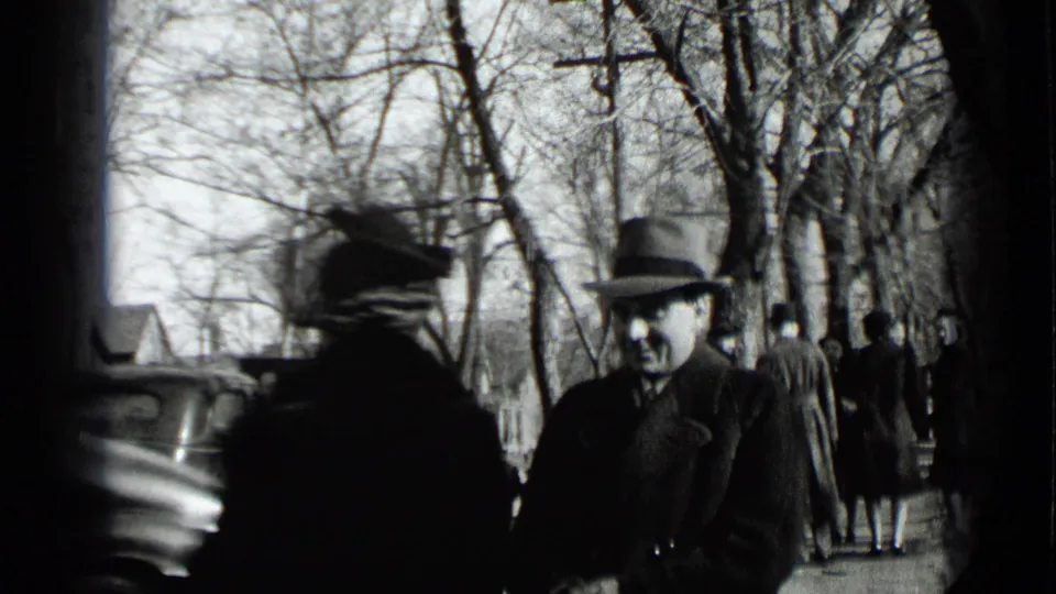 Bundled adults pass along a New York sidewalk with parked cars, bare trees, and winter coats visible in the 1939 home-movie footage.