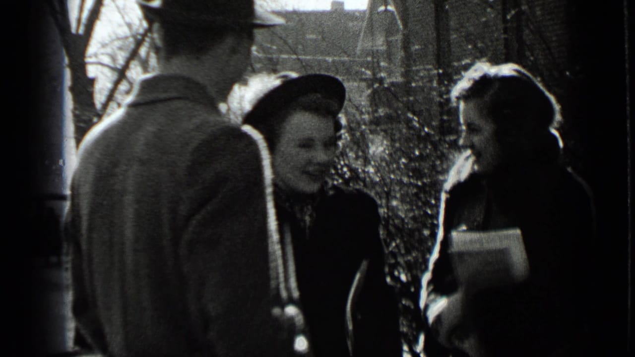 Adults in winter coats and hats talk on a sunlit sidewalk.
