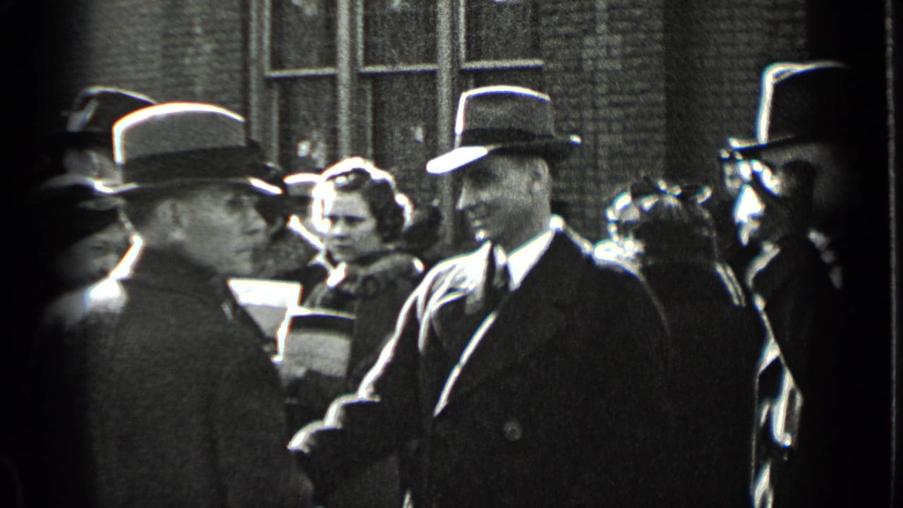 Formally dressed adults stand outside a brick building as a suited man in a fedora faces the camera.