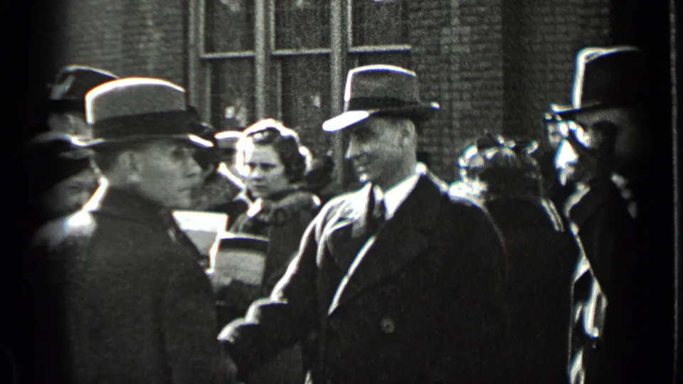 In 1939 New York, a suited man in a fedora smiles toward the camera among formally dressed adults outside a brick building.