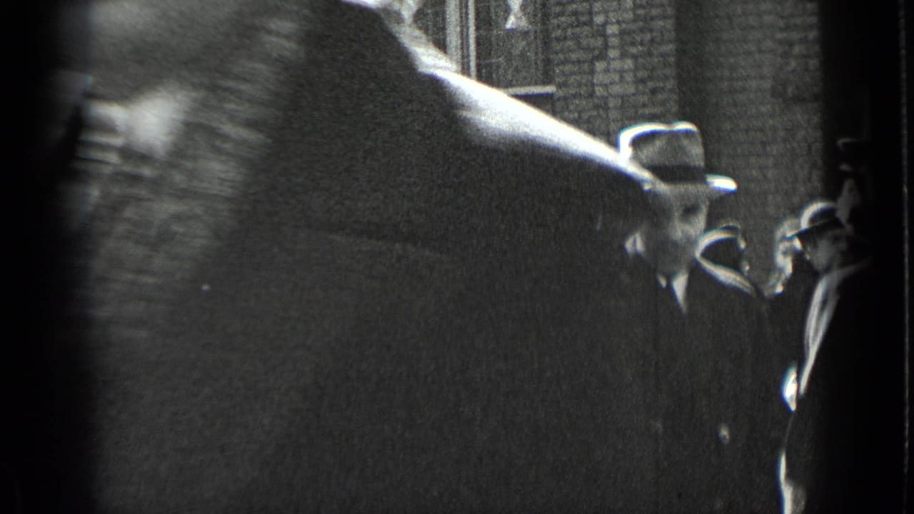 A blurred close figure crosses the frame, partly blocking adults in hats near a brick wall.