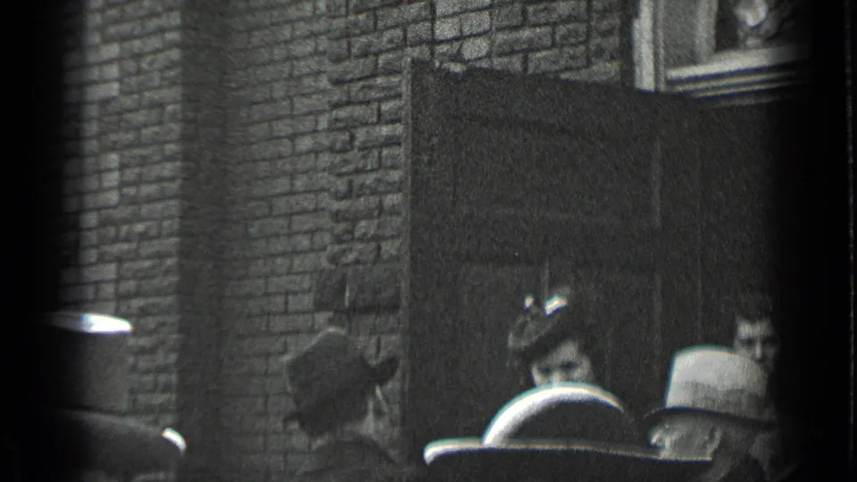 Formally dressed adults cluster near a doorway in a brick New York exterior, with hats and faces partly visible.