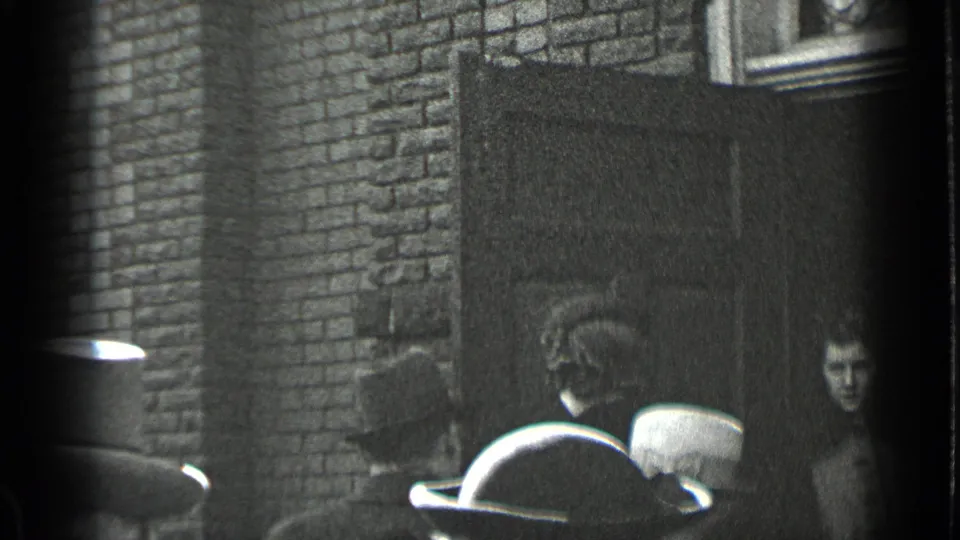 The crowd remains near the brick doorway in 1939 New York, with brimmed hats in the foreground and a face at the edge.