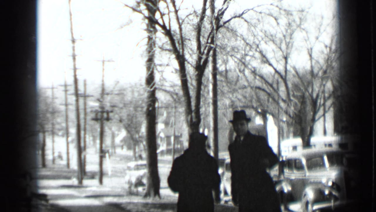 Two adults in coats and hats stand on a snowy sidewalk beside bare trees and parked cars.