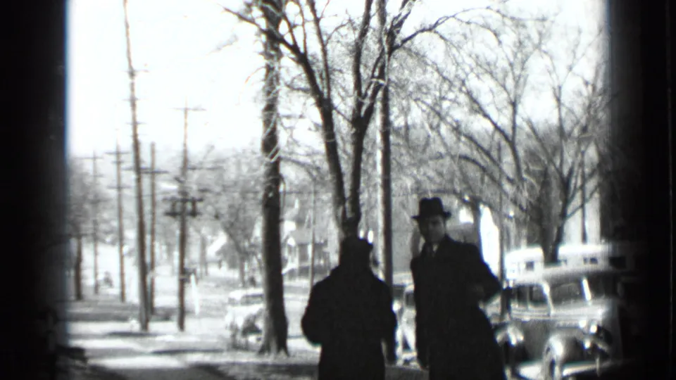 In a 1939 New York 8mm home-movie frame, two adults in dark winter coats and hats stand on a snowy sidewalk lined with bare trees and parked cars.