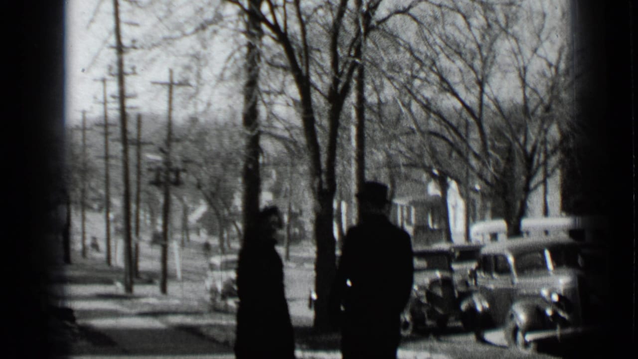 Two adults in winter clothing walk away from the camera on a snowy New York sidewalk, with bare trees, utility poles, and parked cars visible.