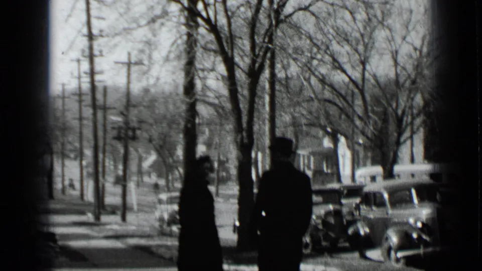 Two adults in winter clothing walk away from the camera on a snowy New York sidewalk, with bare trees, utility poles, and parked cars visible.