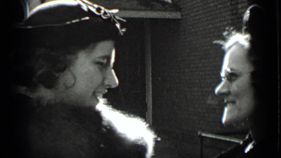 Close 8mm frame of two adults in winter clothing facing each other near a brick wall, with one wearing a decorated hat and fur-trimmed coat.