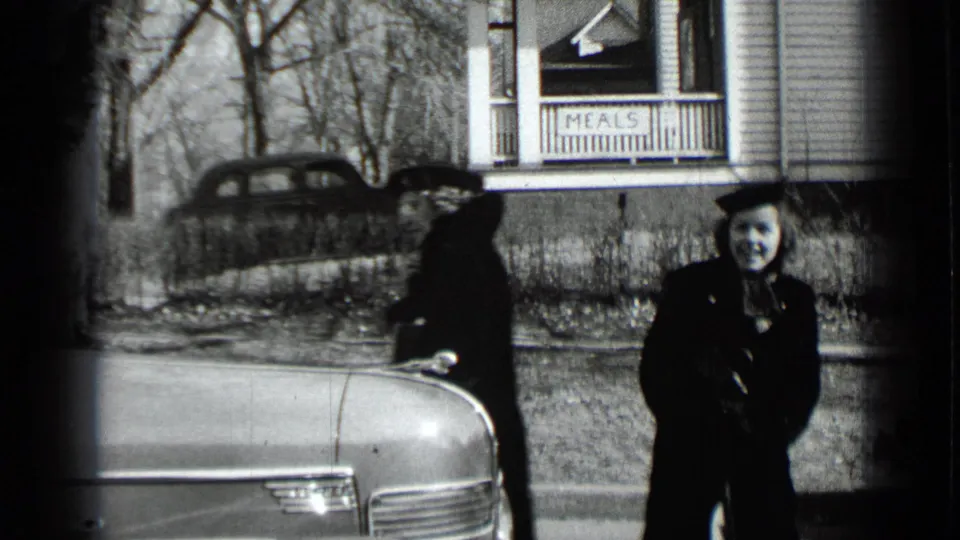 Two women stand beside a rounded 1930s-style car outside a New York roadside house with a visible porch sign reading “MEALS.”