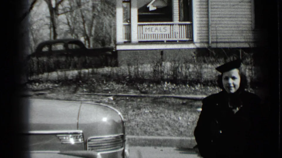 A woman in dark outerwear stands near a rounded car outside a New York house with a porch sign reading “MEALS.”